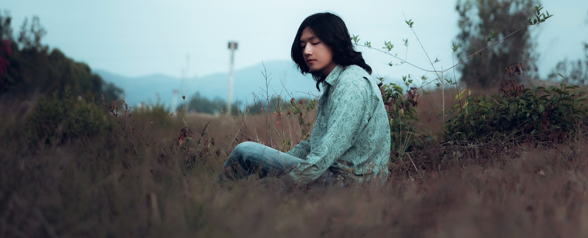 Young man with shoulder-length hair sitting quietly in a grassland